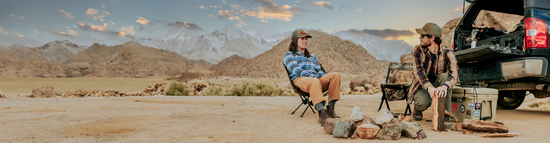 Two people sitting in chairs by a truck in a desert landscape with mountains in the background.