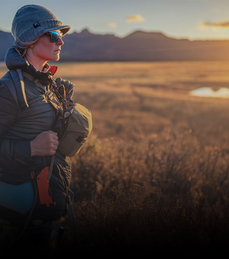 Person standing in a field with mountains in the background during sunset.